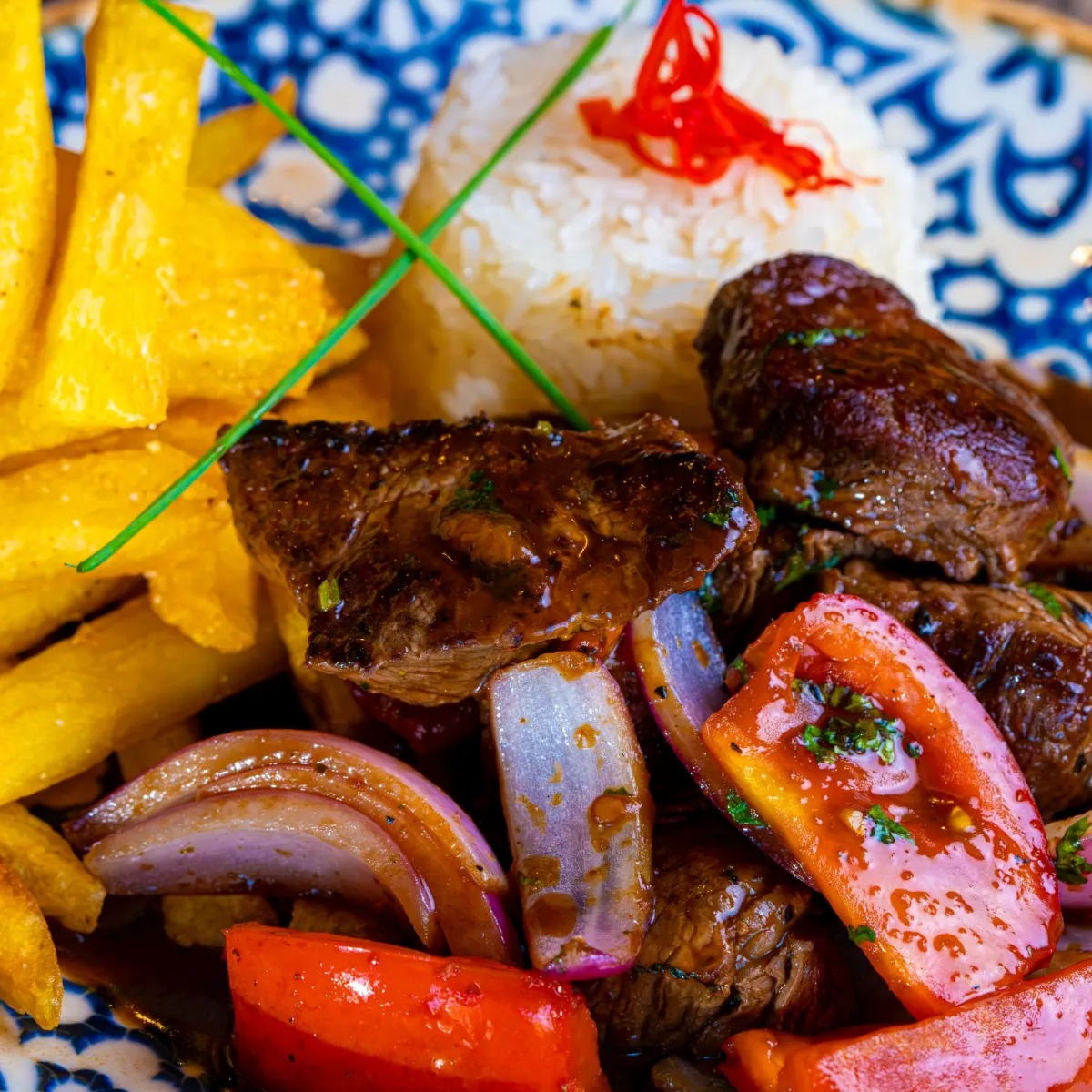 Lomo saltado con tiras de carne, cebolla morada y tomate, papas fritas gruesas y arroz blanco en un plato de cerámica azul